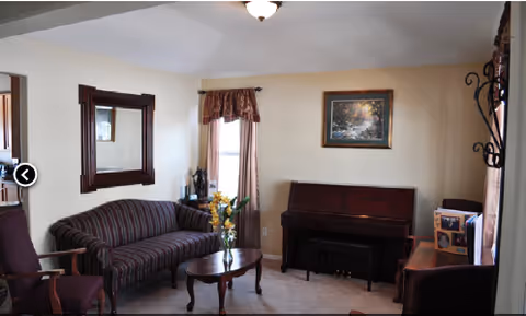 A cozy living room with a striped sofa, a wooden coffee table with a vase of yellow flowers, a piano against the wall, a framed painting above the piano, a large mirror on the wall, and several chairs arranged around the room. The room has beige walls and carpeted floor.