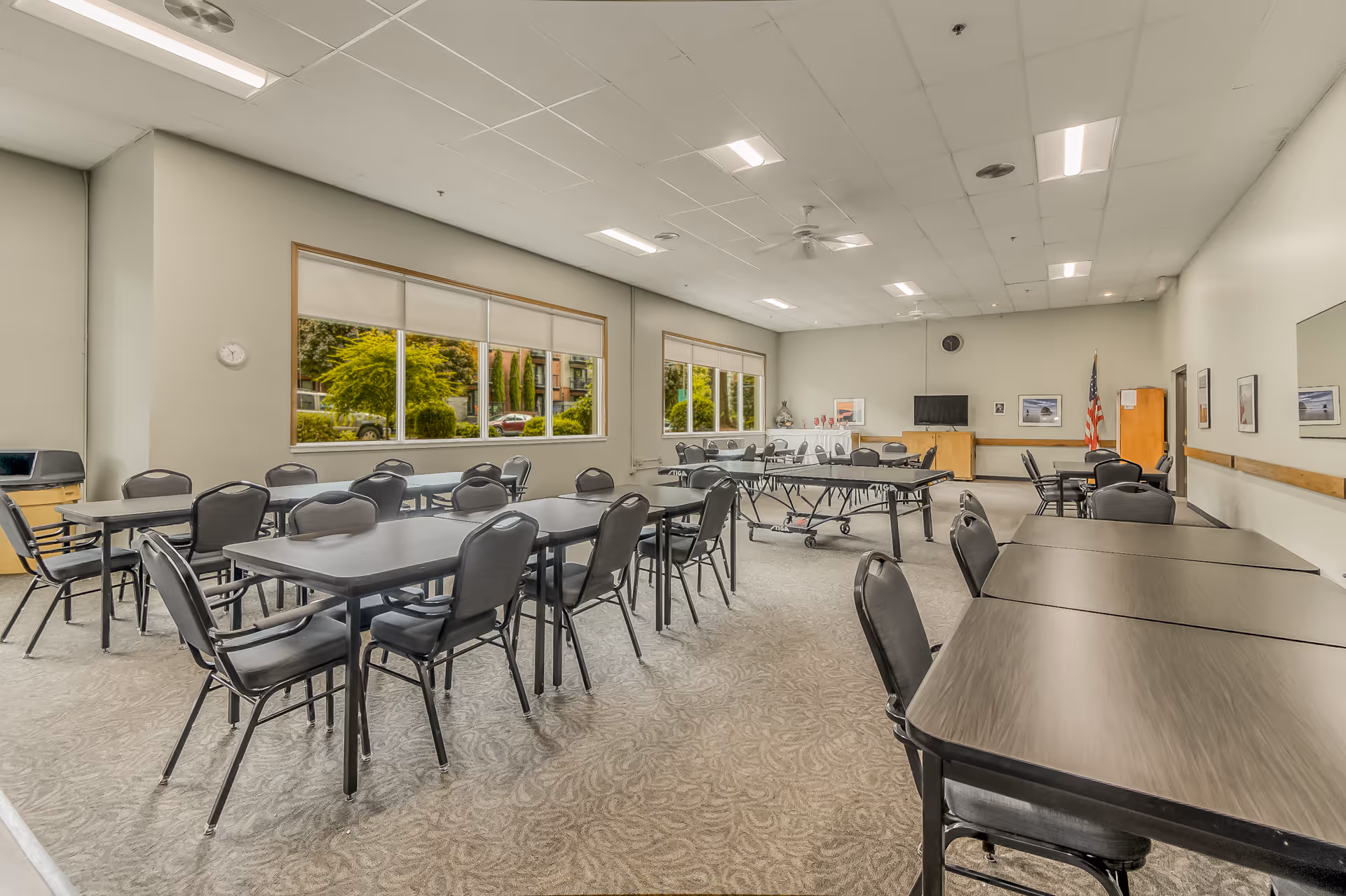 A spacious, well-lit dining room with multiple black tables and chairs arranged neatly. Large windows on one side let in natural light and show greenery outside. The room has a carpeted floor, a ceiling fan, a wall-mounted TV, and an American flag in the corner.