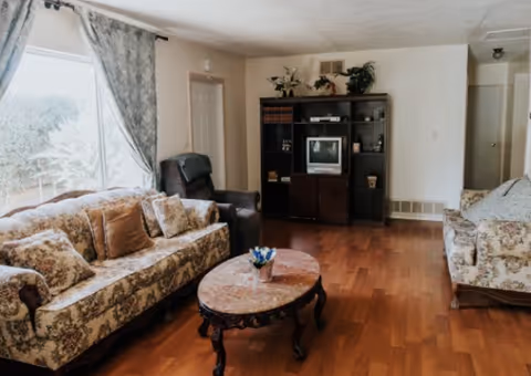 A cozy living room with two floral-patterned sofas, a dark armchair, a round wooden coffee table with a small flower arrangement, a large window with floral curtains, and a dark wooden entertainment center holding a TV and decorative items.