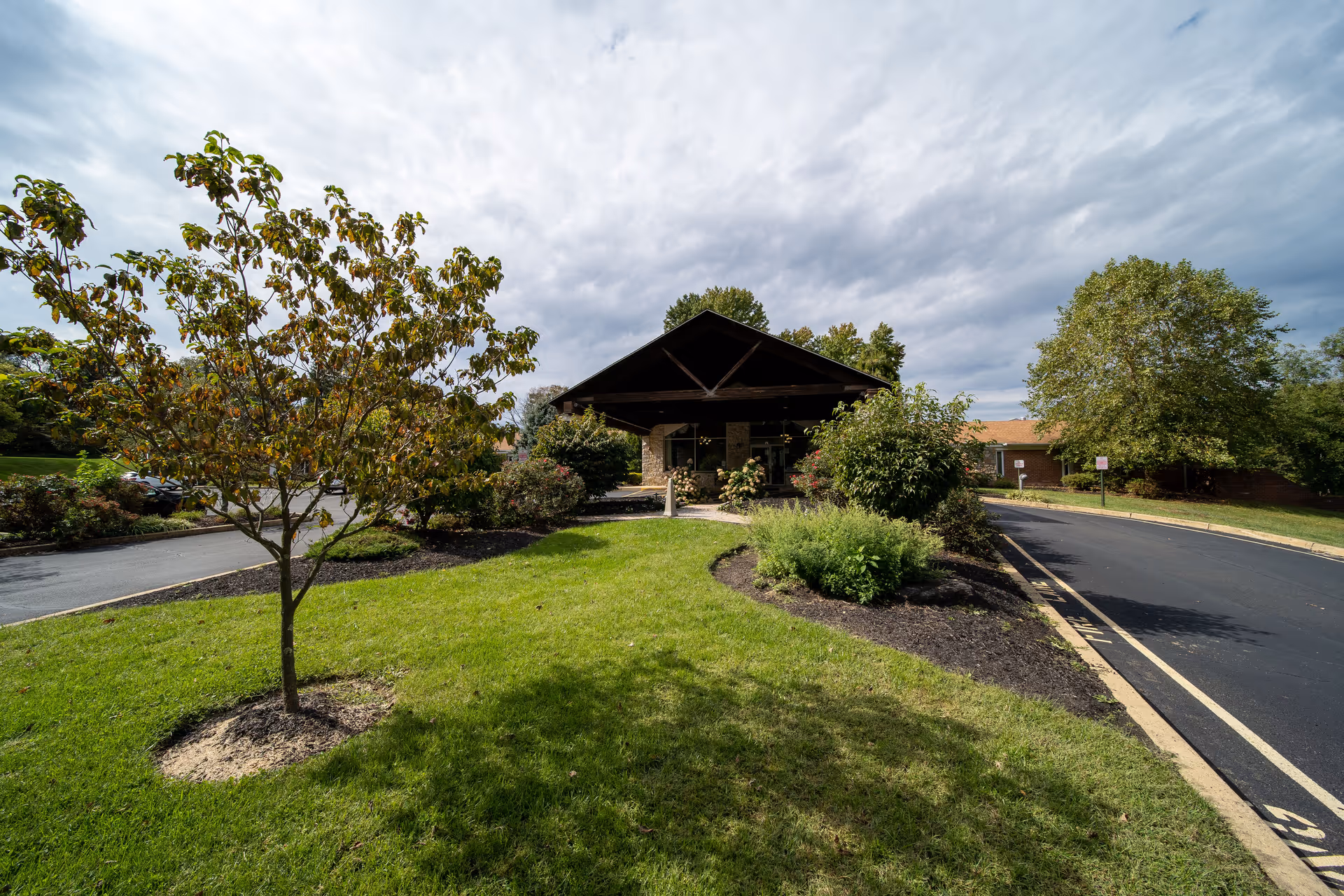 View of the front entrance of Symphony Manor of Feasterville, featuring a covered driveway with a peaked roof, surrounded by green grass, trees, and shrubs under a cloudy sky.
