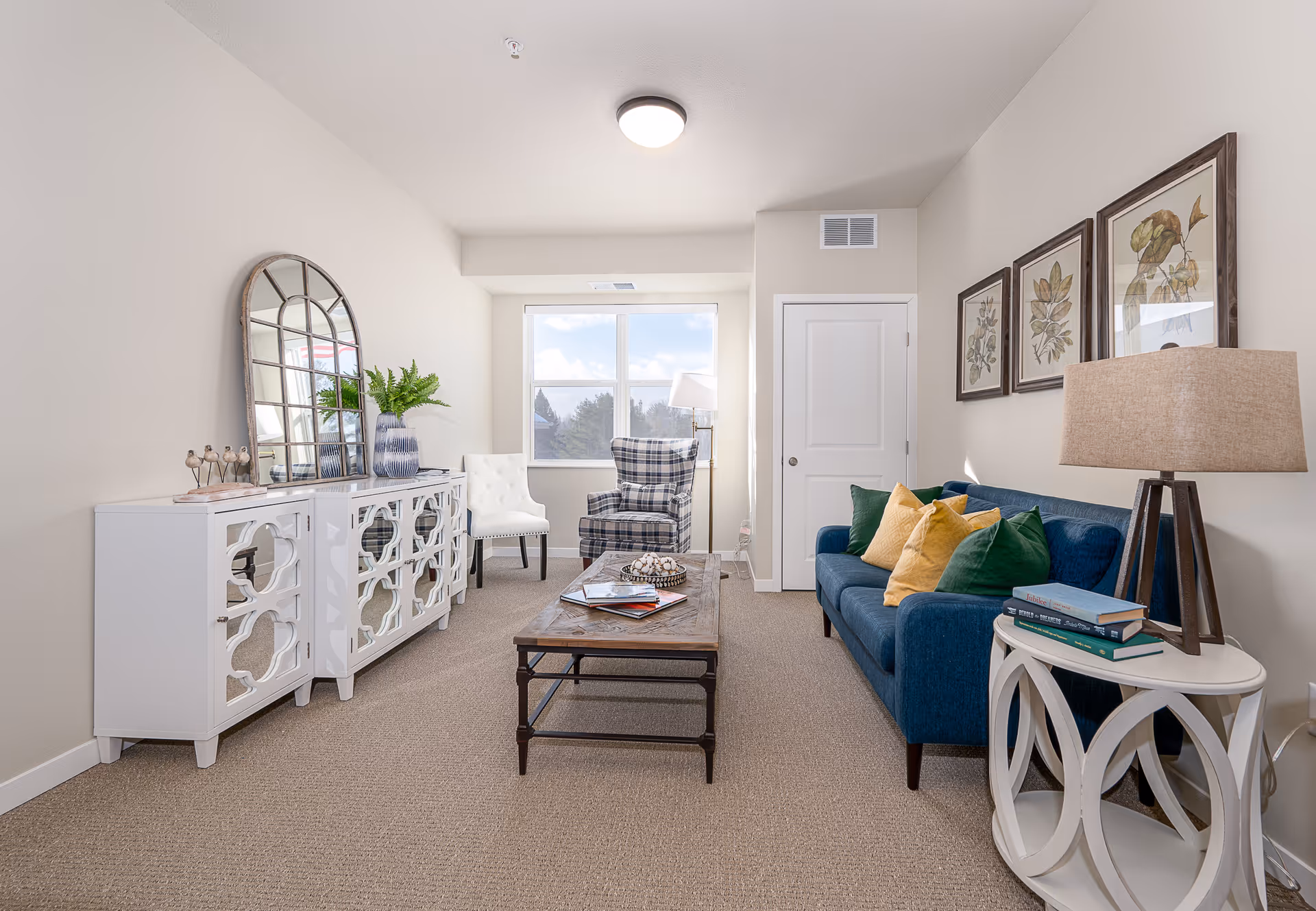 Bright furnished living room with a blue sofa, coffee table, accent chairs, and a decorative console under a window.