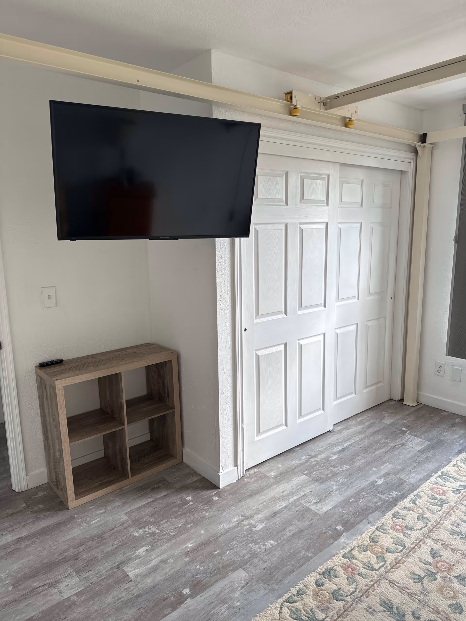 A corner of a room with a wall-mounted flat-screen TV above a small wooden shelf unit with four compartments. Next to the shelf is a white double sliding door closet. The floor has gray wood-like vinyl planks and a floral patterned rug is partially visible on the right side.