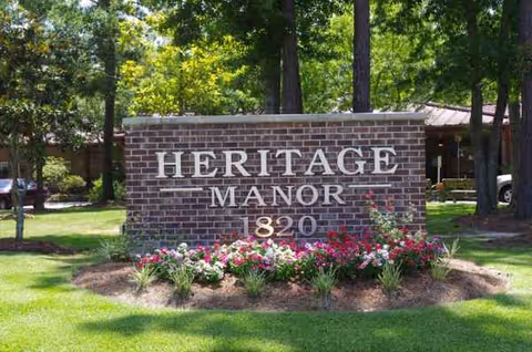 Brick sign reading Heritage Manor with flower beds and trees in front.