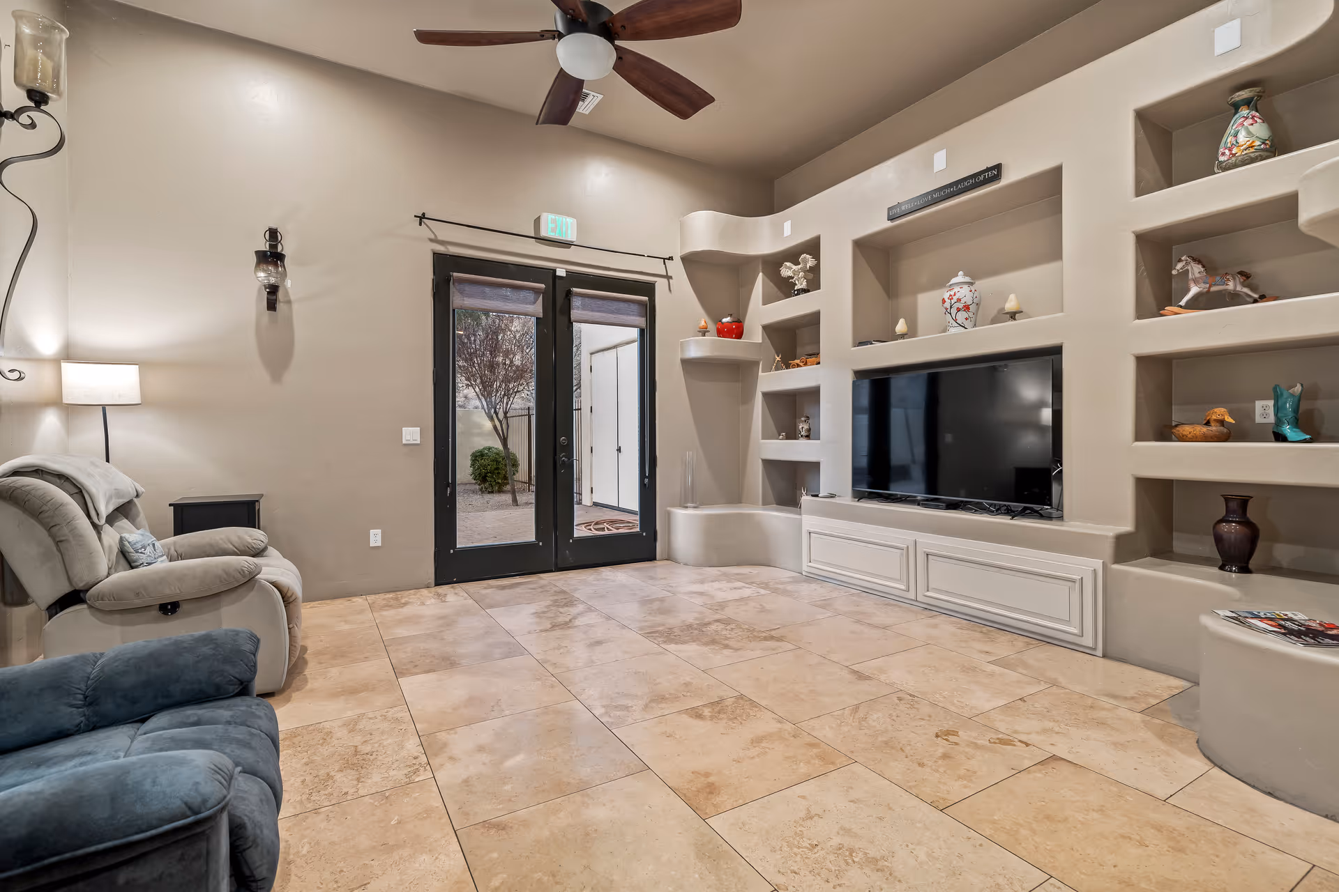 Spacious lounge with tiled floor, built-in shelving and TV, recliner chairs, and double doors leading outside.