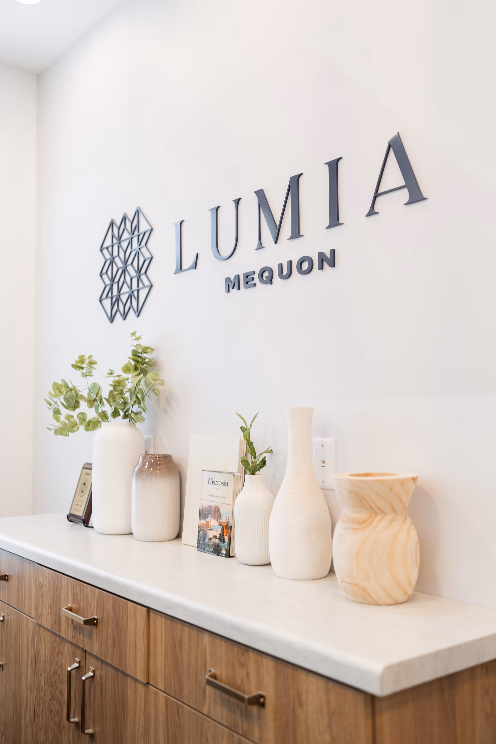 A countertop with decorative vases and a small plant in front of a white wall with the text 'LUMIA MEQUON' and a geometric logo mounted on it.