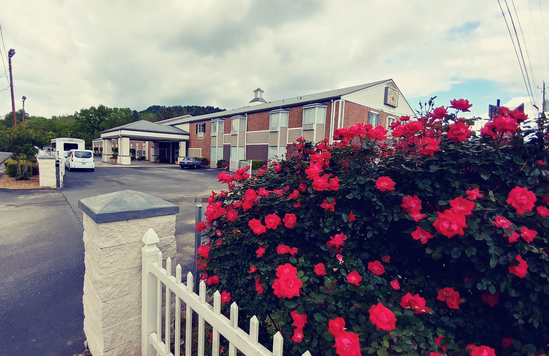Exterior view of Cherry Creek Village, an affordable independent living community for seniors, showing a two-story brick building with a covered entrance, a parking area with vehicles, and vibrant red flowers in the foreground near a white fence.