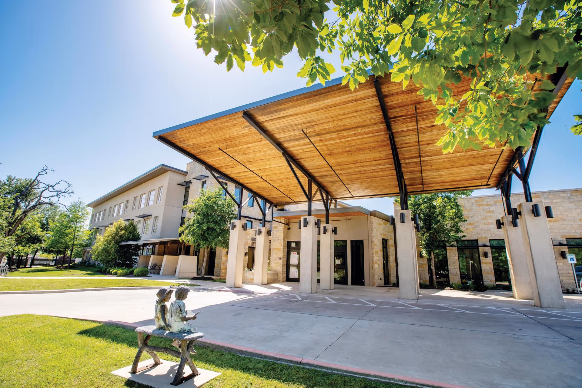 Front entrance of a senior living building with a large wooden canopy supported by stone columns and a small bronze bench statue on the lawn.