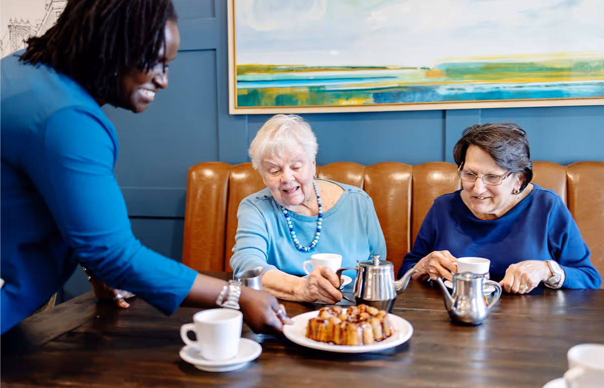A smiling woman serves a plate of pastries to two elderly women sitting at a table with cups of tea or coffee in a cozy dining area with a leather bench and a colorful abstract painting on the wall.