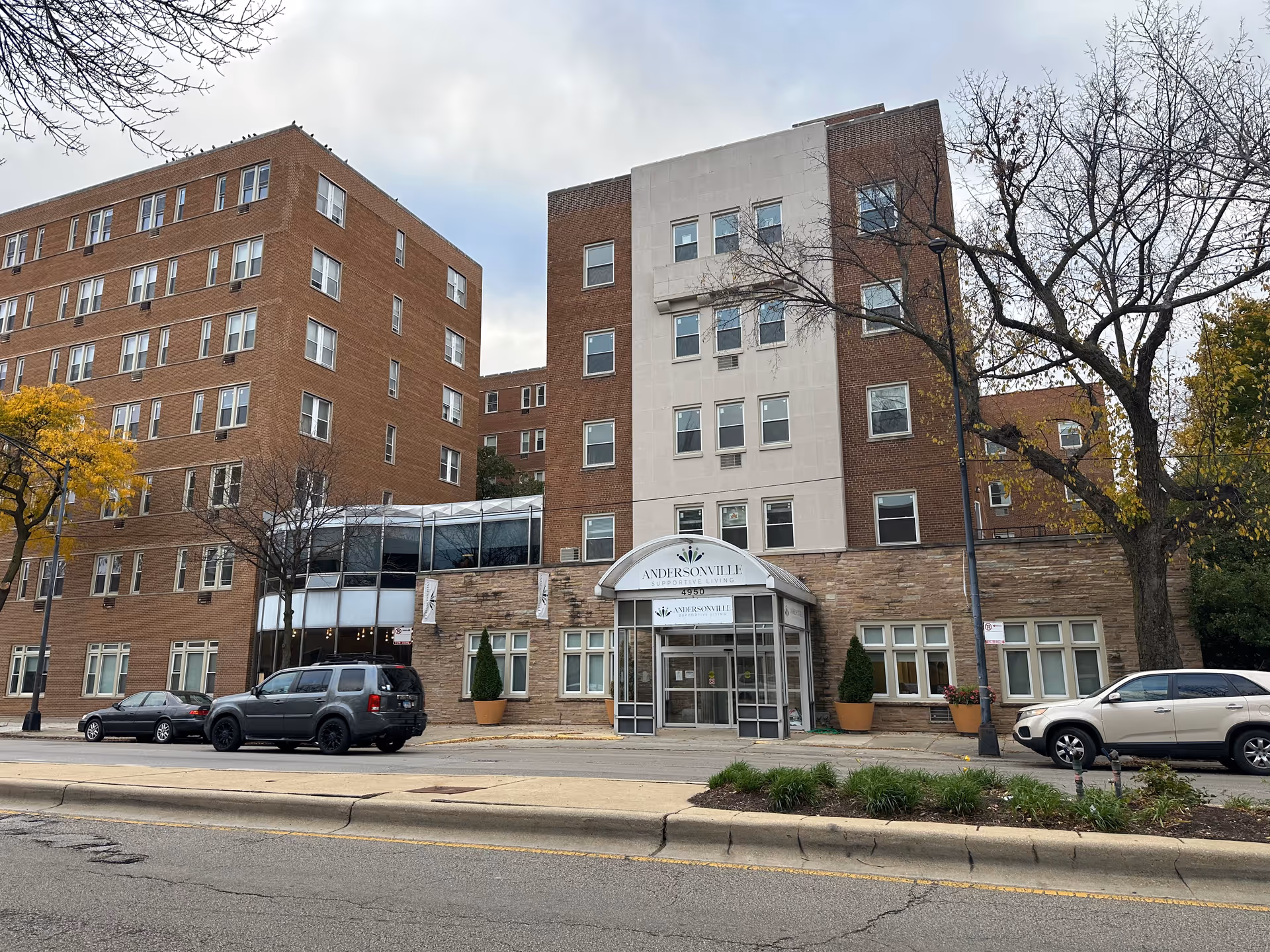 Front exterior of a multi-story brick retirement community building with an entrance canopy and cars parked on the street.
