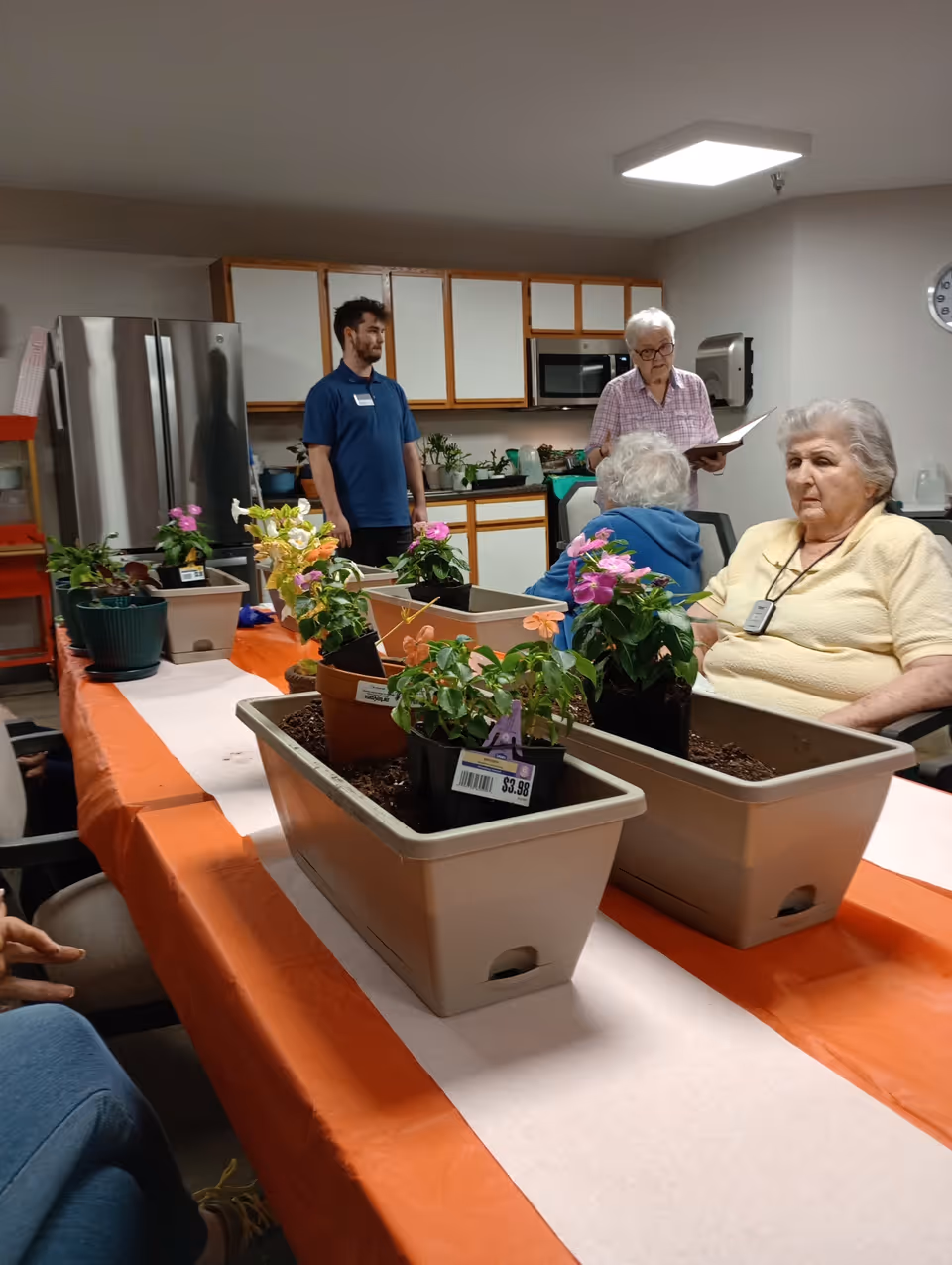 Several seniors and a staff member sit around a long table with potted flowers and planters in a communal activity room with a kitchenette.