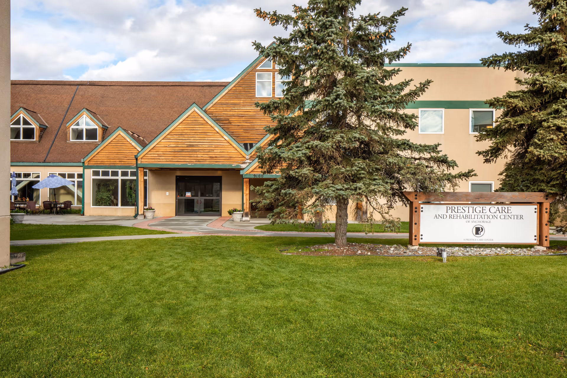 Front entrance of a post-acute care building with a grassy lawn, evergreen trees, and a sign for Prestige Care and Rehabilitation Center.