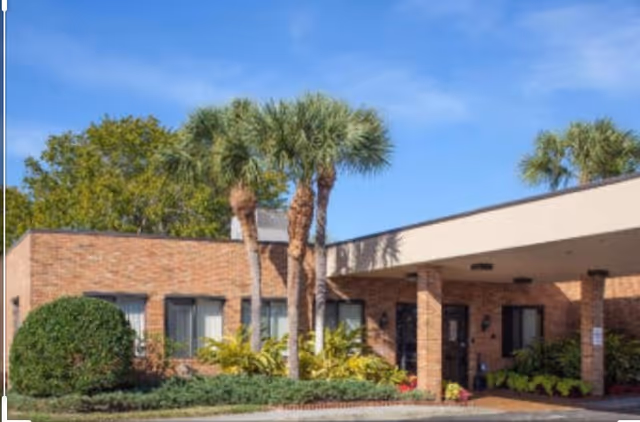 Exterior view of a single-story brick building with a covered entrance, surrounded by palm trees and shrubs under a clear blue sky.