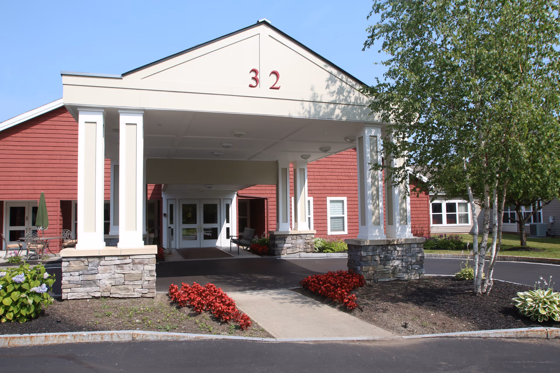Entrance of a senior living facility with a covered drop-off area supported by white columns with stone bases. The building has red siding and white trim, with the number 32 displayed at the top of the entrance. There are flower beds with red flowers and green shrubs on either side of the walkway leading to the double glass doors. Trees and additional parts of the building are visible in the background under a clear blue sky.