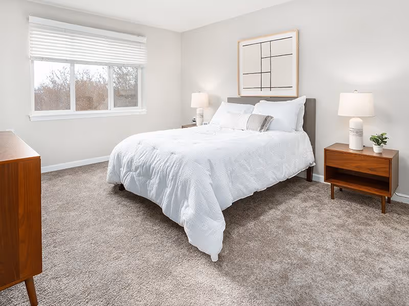 A bright and clean bedroom with a large window on the left wall. The room features a bed with white bedding and pillows, flanked by two wooden nightstands each with a white lamp. There is a wooden dresser on the left side of the room and a modern abstract framed artwork hanging above the bed. The floor is carpeted in a light brown color.