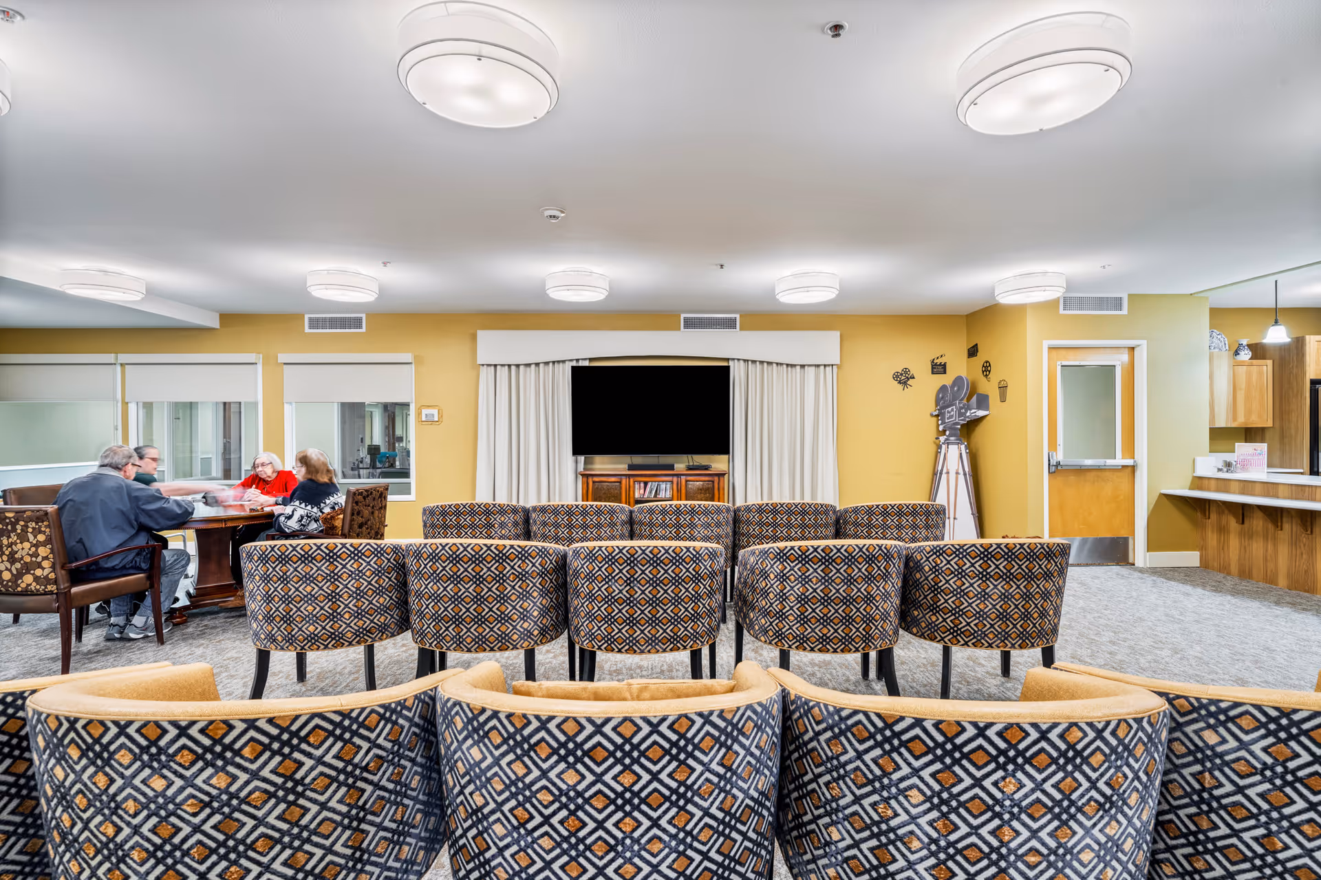 A common area in a senior living facility with patterned chairs arranged in rows facing a television mounted on a yellow wall. To the left, four elderly people are seated around a table engaged in conversation. The room has bright ceiling lights, windows with white blinds, and a decorative vintage film camera on a tripod near a door. A kitchen counter with wooden cabinets is visible on the right side.
