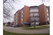 Exterior view of a multi-story brick building with large windows and a blue awning over the entrance, surrounded by a paved driveway and some leafless trees on a cloudy day.