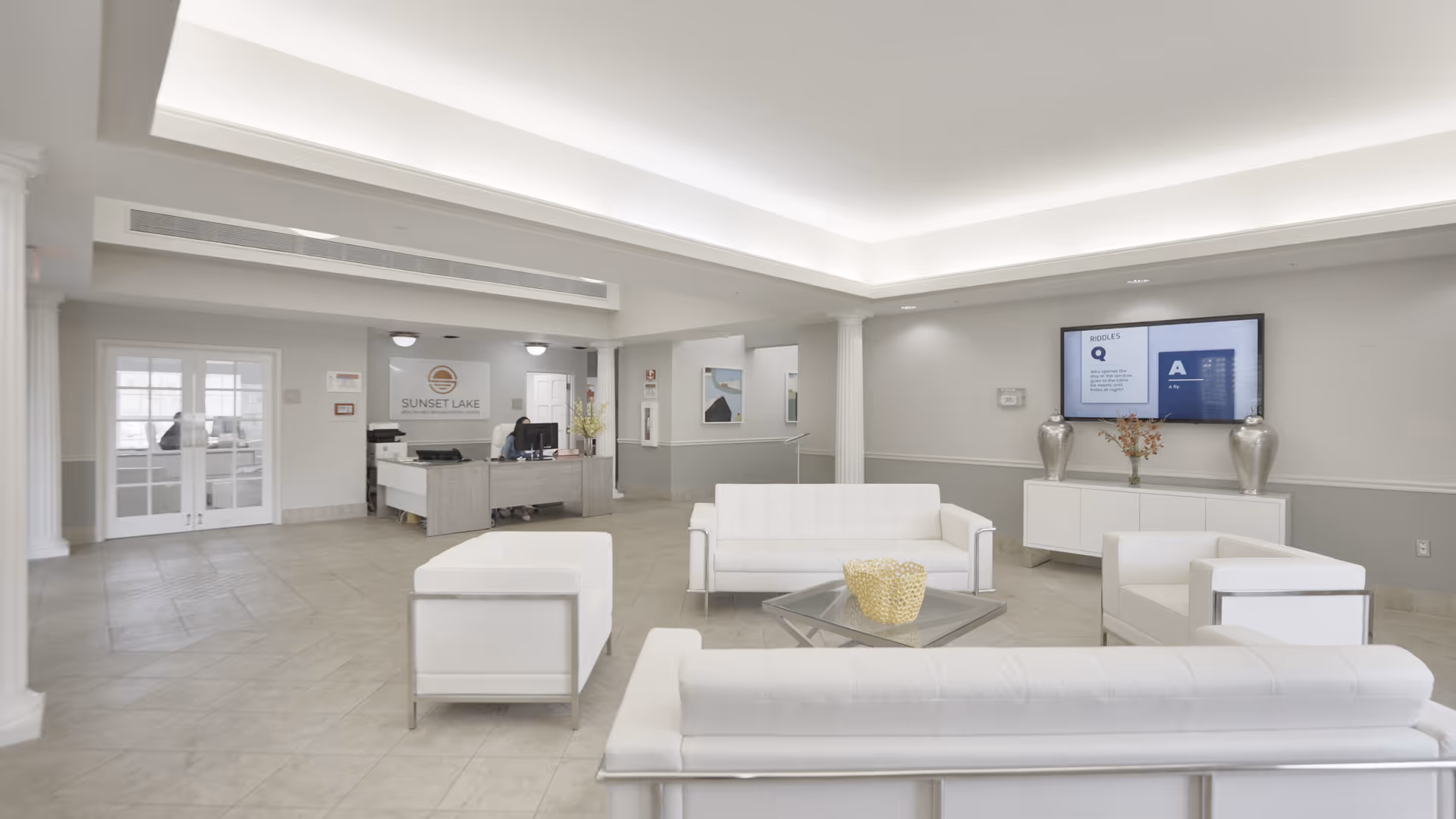 A bright and spacious reception area of Sunset Lake Health and Rehabilitation Center featuring modern white sofas and armchairs arranged around a glass coffee table. A receptionist is seated at a desk in the background with the facility's logo on the wall behind. The room has light-colored tiled flooring, neutral walls, and a large flat-screen TV mounted on the wall displaying information.