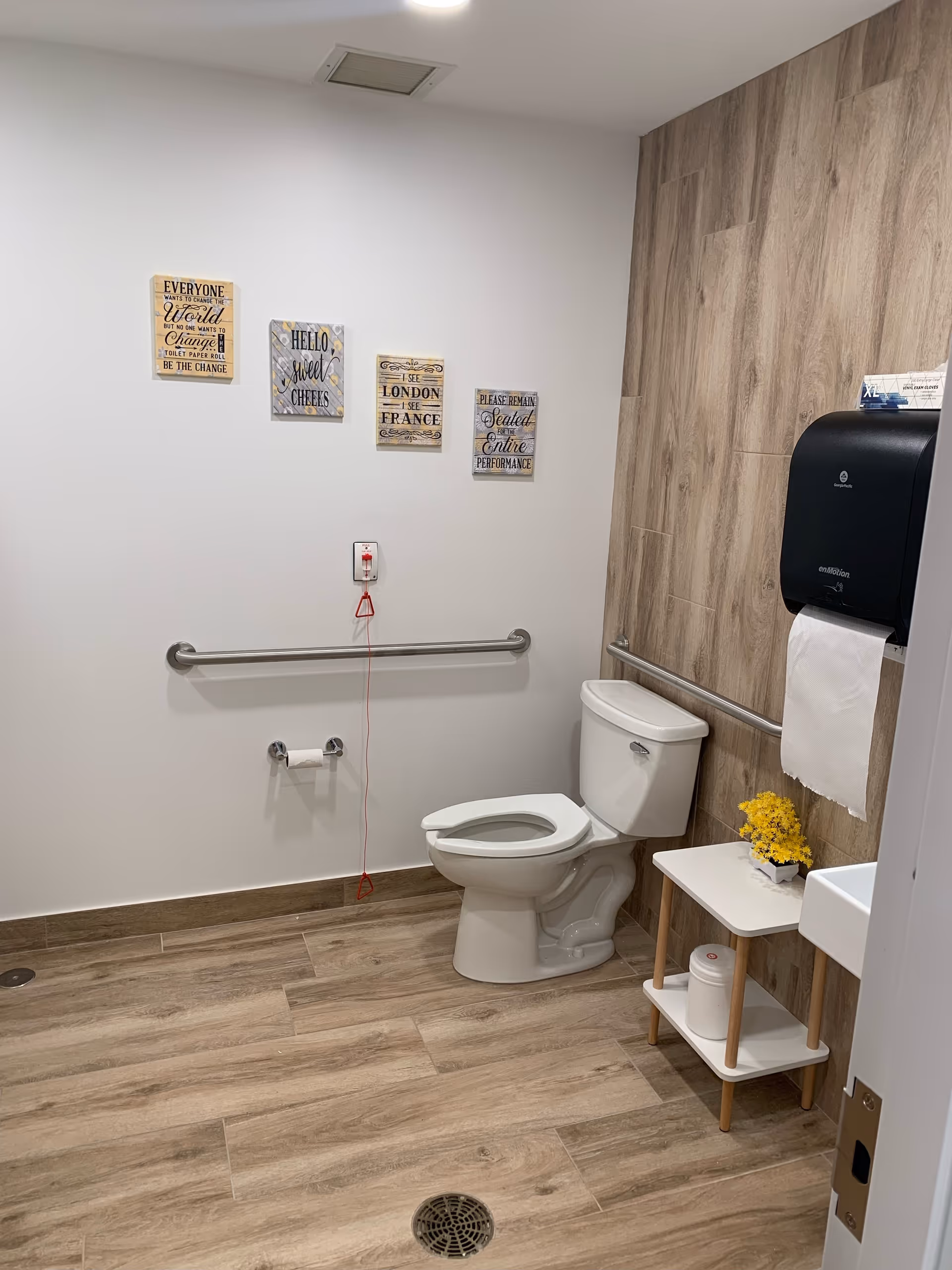 A clean and accessible bathroom with a white toilet, grab bars on the walls, a small white table with yellow flowers and a container underneath, a paper towel dispenser, and four decorative wall signs with various quotes. The floor and one wall have wood-like tile, and the other walls are white.