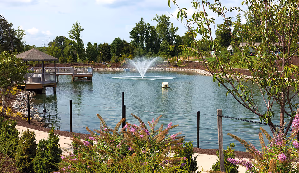 A landscaped pond with a central fountain, gazebo-covered docks, walkways, and flowering shrubs.