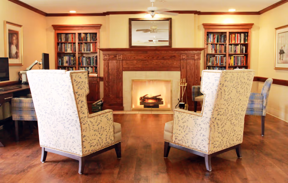 A cozy living room area with a wooden floor, two patterned armchairs facing a lit fireplace with a wooden mantel. On either side of the fireplace are built-in bookshelves filled with books. There is a mirror above the fireplace reflecting ceiling fans, and additional seating and framed artwork are visible on the walls.