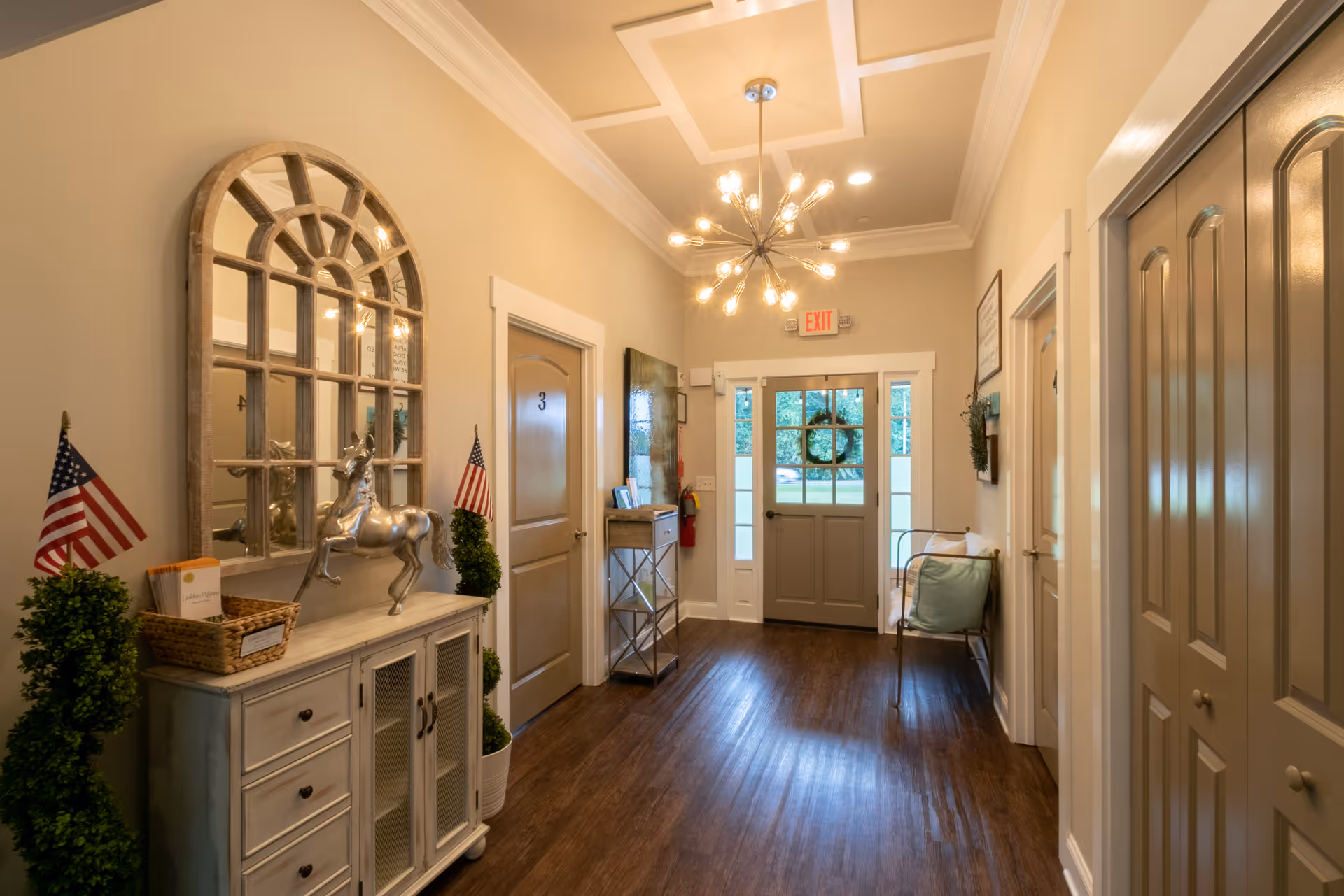 A well-lit hallway in a senior living facility with wooden floors, beige walls, and white trim. The hallway features a decorative cabinet with a silver horse statue, two small American flags, and a large arched mirror above it. There is a door labeled with the number 3, a small table with framed pictures, a bench with cushions, and a door at the end with a wreath hanging on it. A modern chandelier hangs from the ceiling.