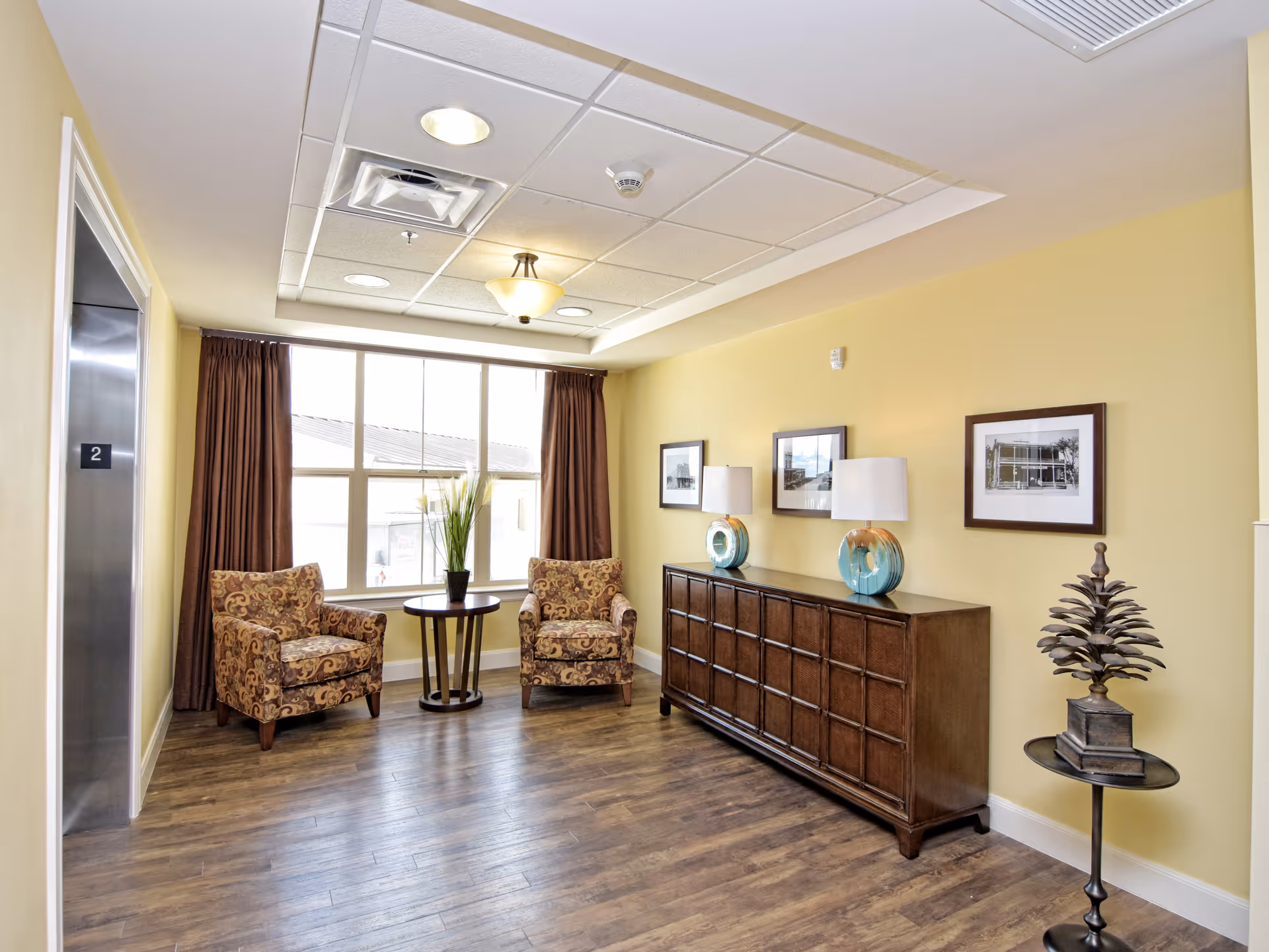A bright interior seating area with two patterned armchairs and a small round table with a plant between them, positioned in front of a large window with brown curtains. To the right, there is a long wooden sideboard with two decorative lamps and framed black and white photos on the yellow wall above it. On the far right, a small round table holds a decorative sculpture. The floor is wood, and an elevator door marked with the number 2 is visible on the left side.