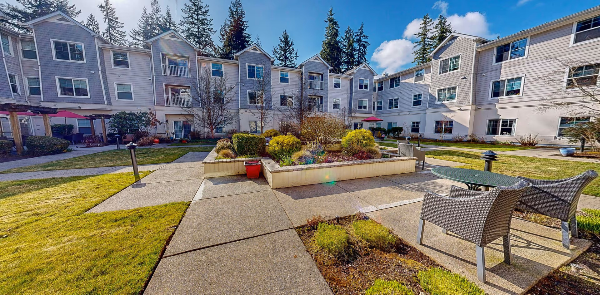Outdoor courtyard area of a senior living facility with a central raised garden bed surrounded by paved walkways, lawn, and patio furniture including a round table and chairs. The three-story building with multiple windows and balconies surrounds the courtyard, with tall evergreen trees in the background under a blue sky with some clouds.
