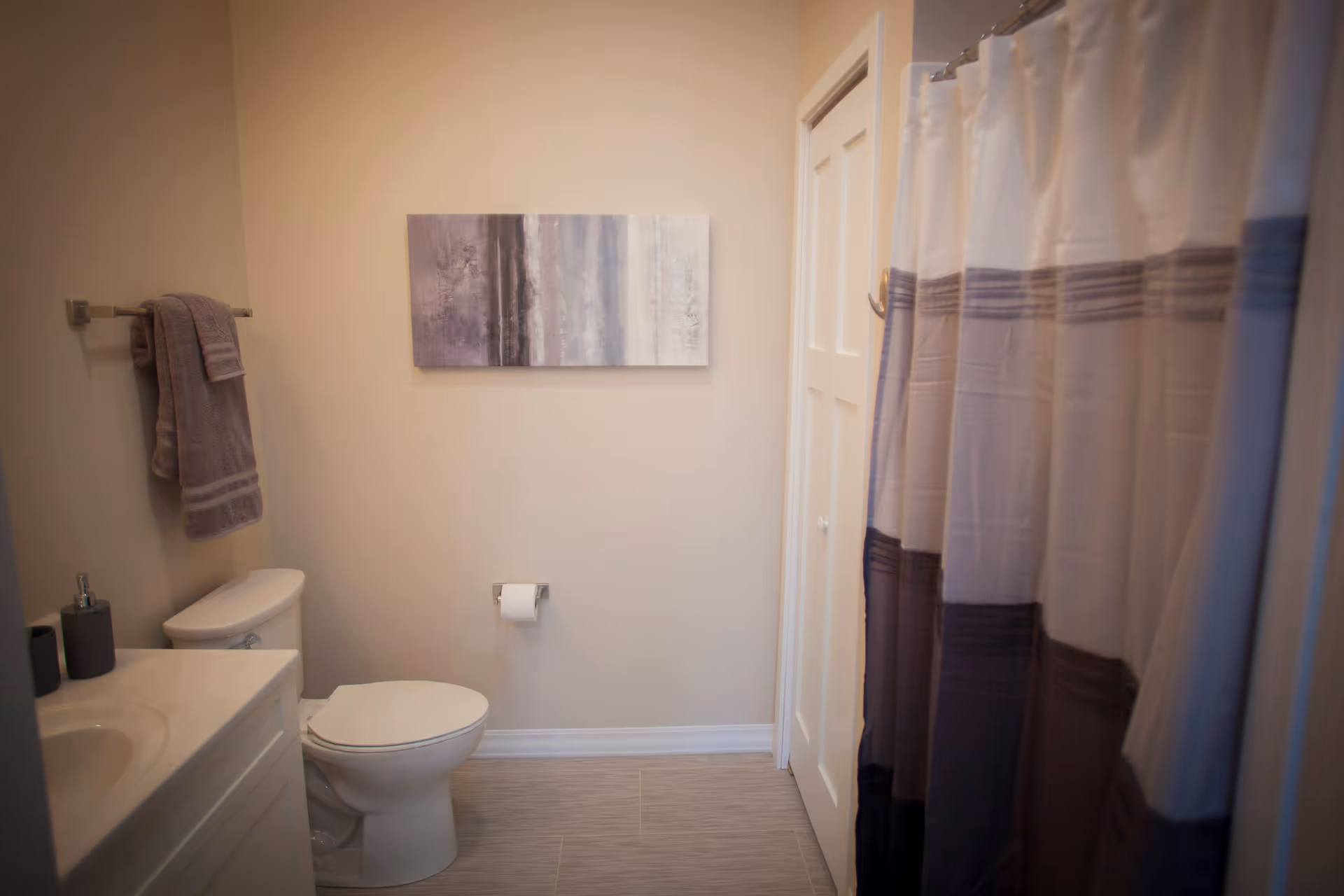 A clean bathroom with a white toilet, a white vanity with a sink, a towel rack holding two gray towels, a piece of abstract wall art above the toilet, and a shower with a curtain featuring horizontal stripes in shades of gray and white.