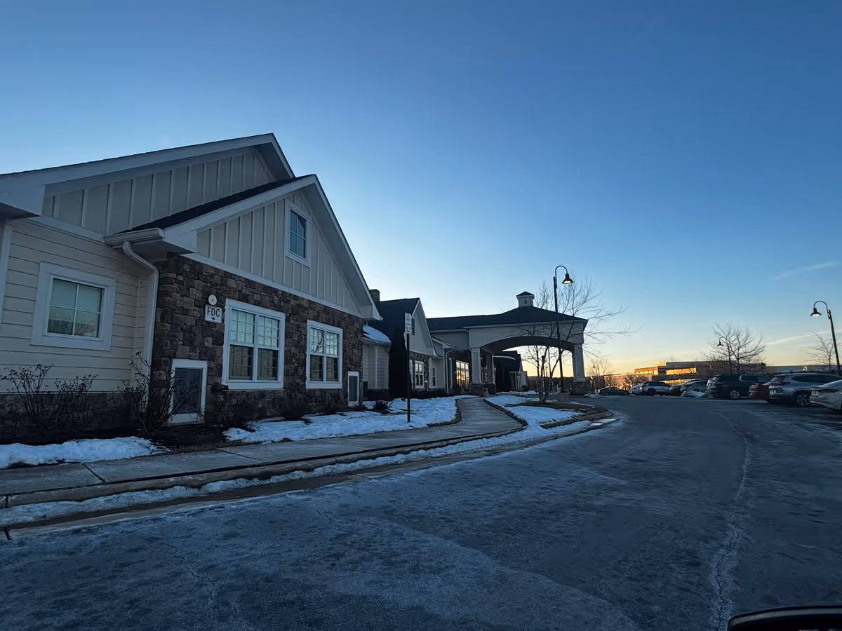 Front exterior of a memory care building with a covered entrance, sidewalk, parked cars and light snow on the ground.
