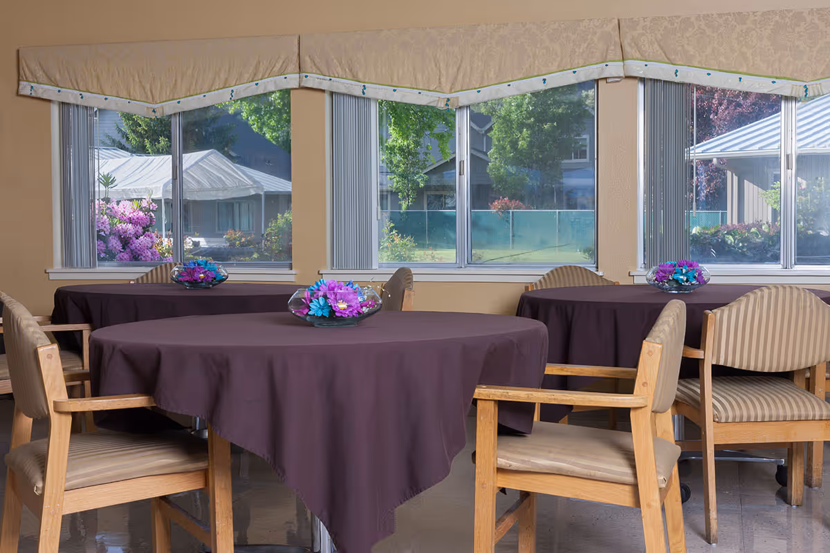 A dining area with round tables covered in dark purple tablecloths, each adorned with a glass bowl containing colorful artificial flowers. Wooden chairs with beige cushions surround the tables. Large windows in the background reveal a view of greenery and buildings outside.