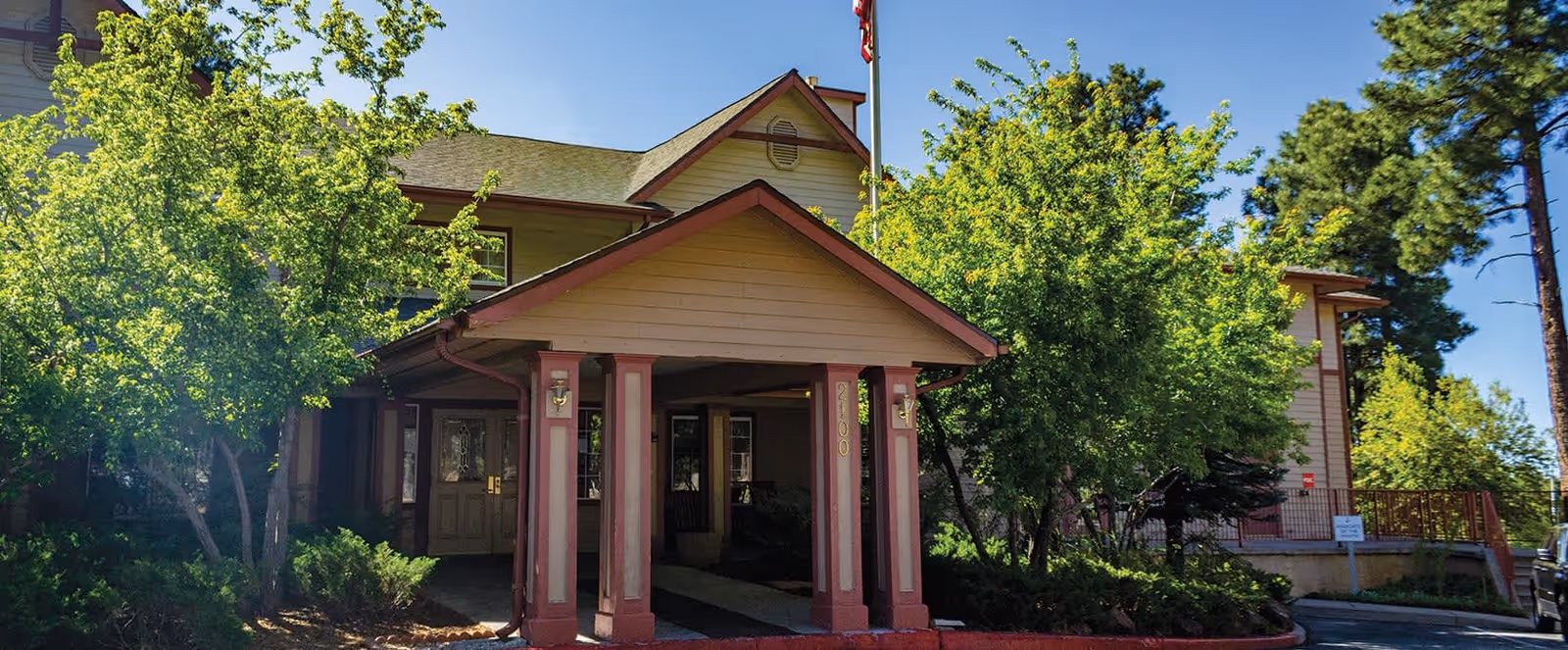 Exterior view of a senior living facility building with a covered entrance supported by four columns. The building is surrounded by green trees and bushes under a clear blue sky.