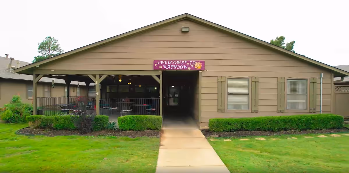 Exterior view of a single-story building with a covered porch area and a sign that reads 'Welcome to Rainbow'. The building is surrounded by green grass and trimmed bushes.