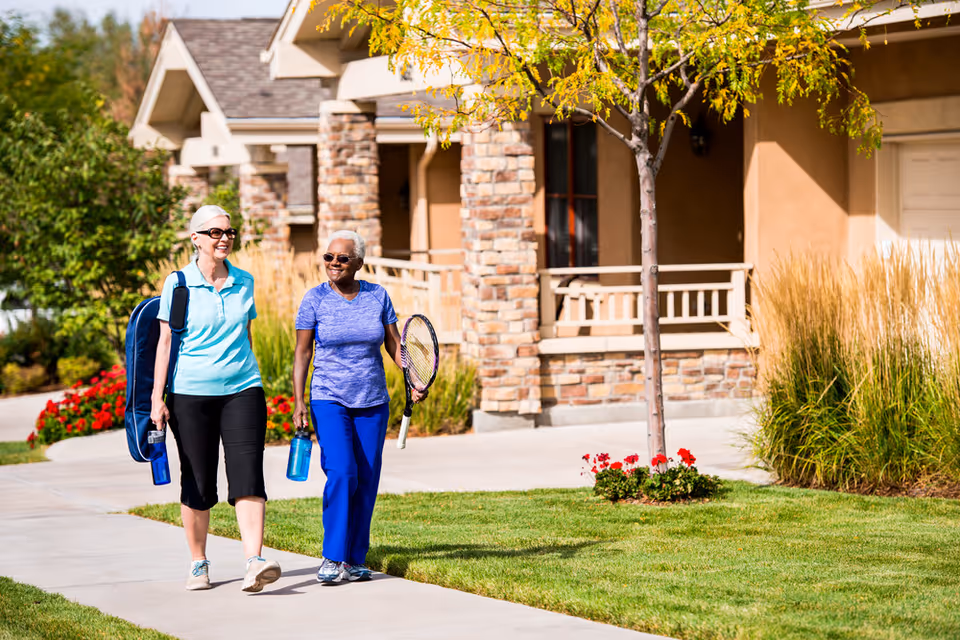 Two elderly women walking on a sidewalk outside a residential building. One woman is carrying a tennis racket and a water bottle, while the other carries a tennis bag and a water bottle. They are smiling and appear to be enjoying a sunny day with greenery and flowers around.