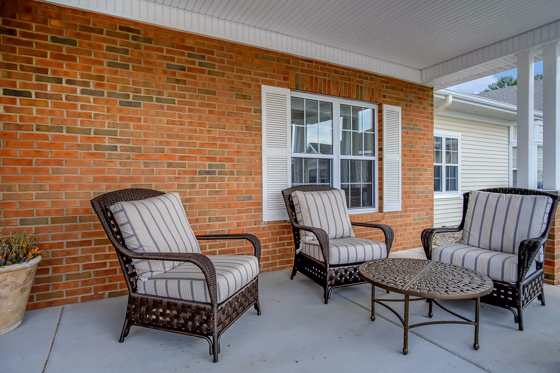 A covered outdoor patio area with three cushioned wicker chairs arranged around a round metal table. The background features a brick wall with a window framed by white shutters, and part of a beige siding exterior wall is visible on the right.