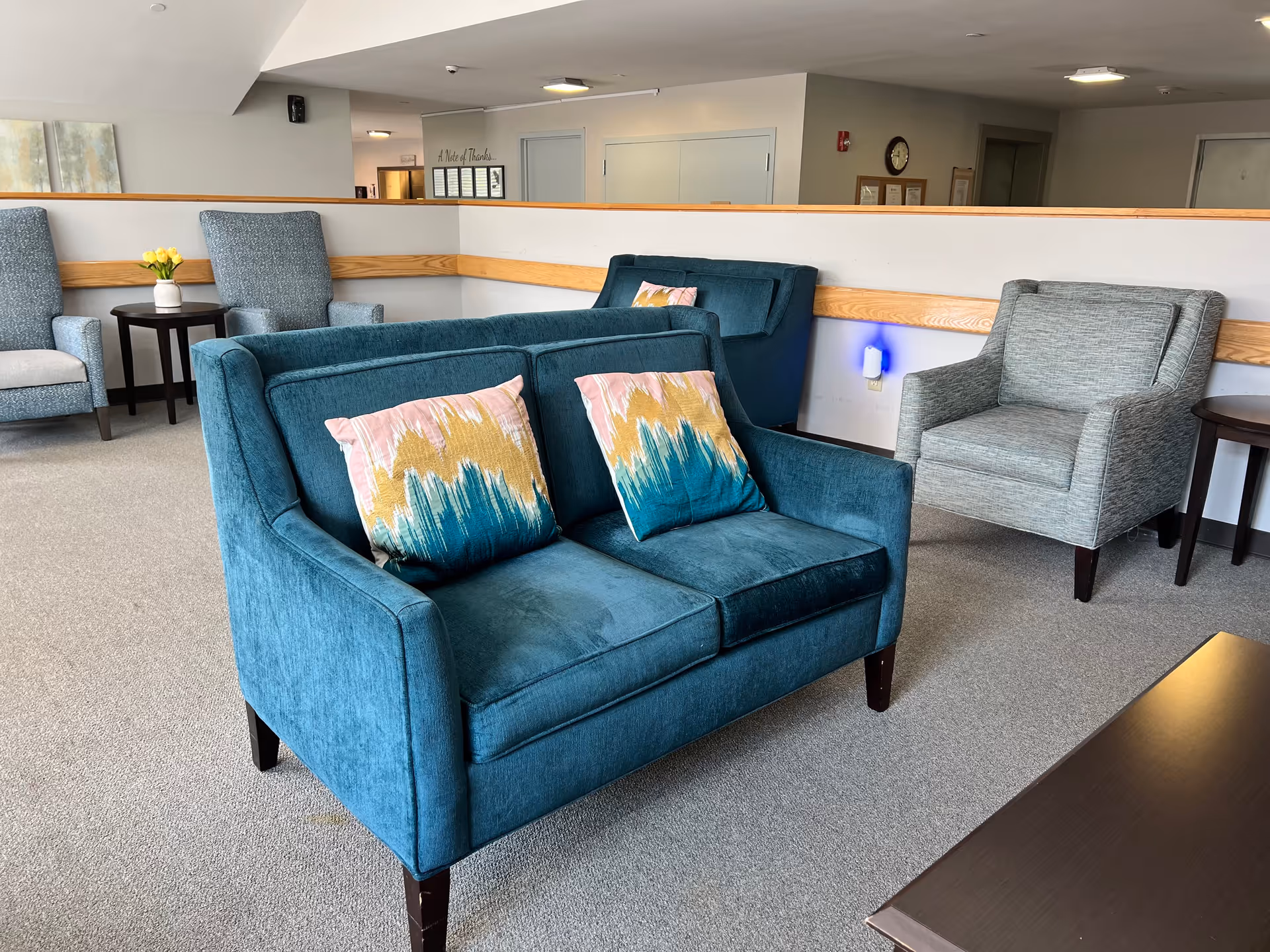 A seating area in a senior living facility featuring a teal loveseat with two colorful patterned pillows, a gray armchair, and another teal loveseat in the background. The room has light gray carpet, light-colored walls with wooden trim, and small dark wood side tables. A vase with yellow flowers is on one of the side tables.