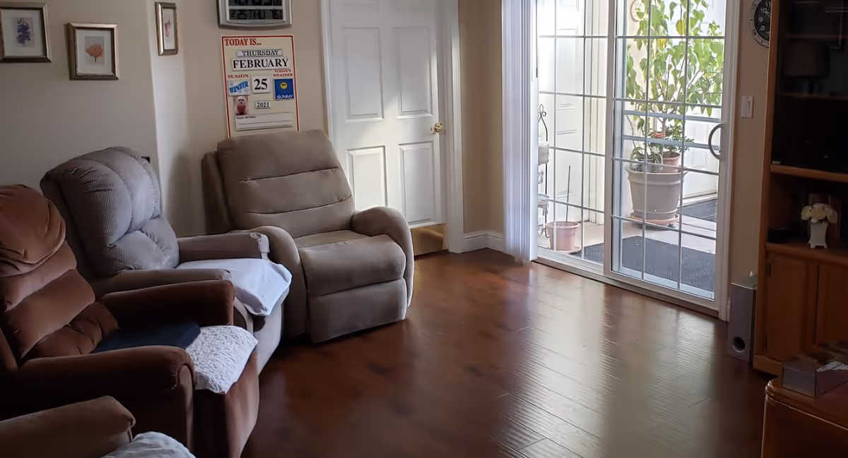 A cozy living room with three recliner chairs in shades of brown and beige, a wooden floor, a sliding glass door leading to an outdoor area with potted plants, and a wall calendar showing the date Thursday, February 25, 2021.