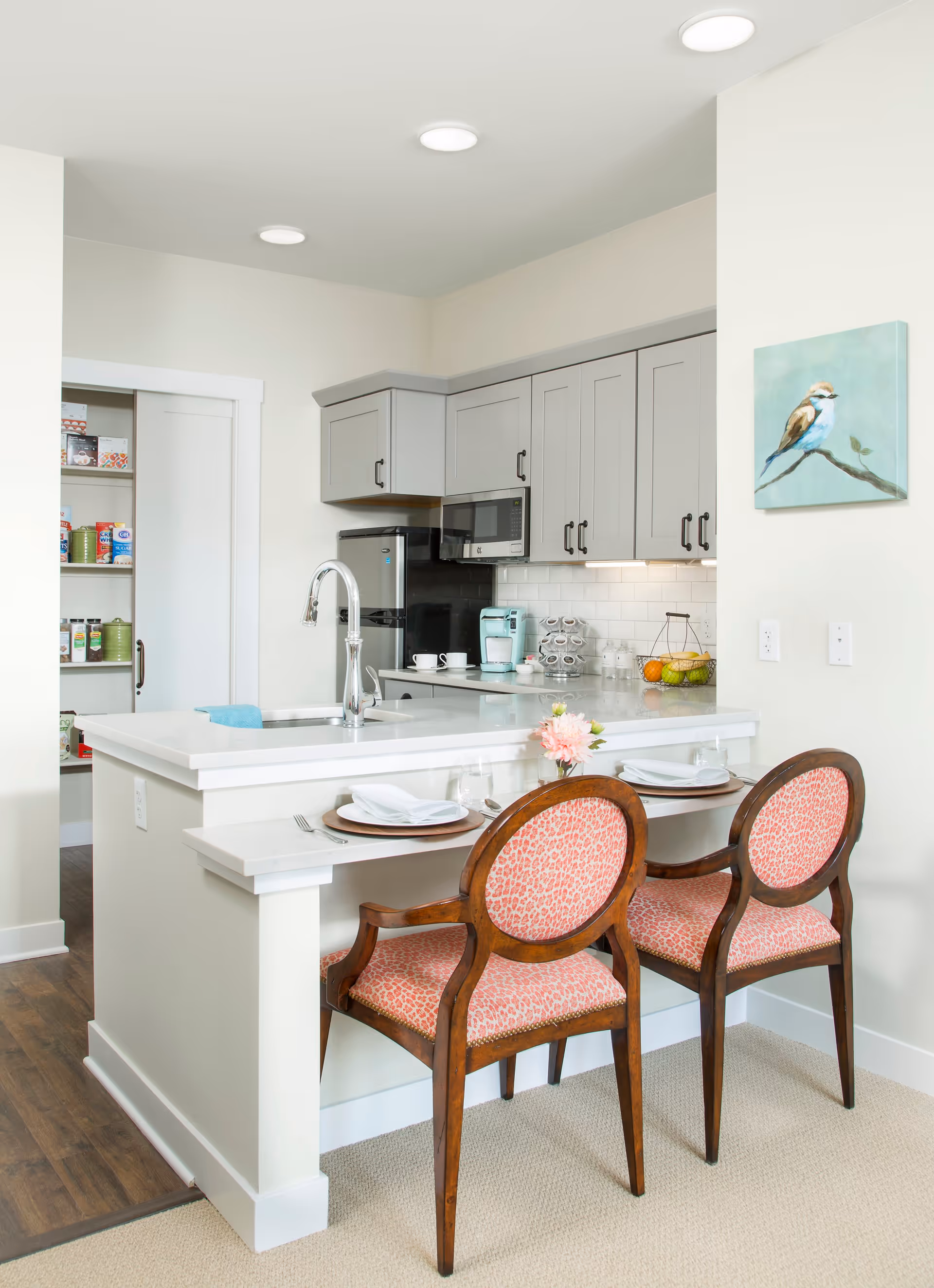 A modern kitchen with light gray cabinets, a white countertop island with a built-in sink and faucet, and two wooden chairs with pink patterned upholstery. The kitchen features a microwave, a small refrigerator, a coffee maker, and a fruit basket. There is a pantry with shelves stocked with food items visible through a sliding door. A painting of a bird on a branch hangs on the wall.