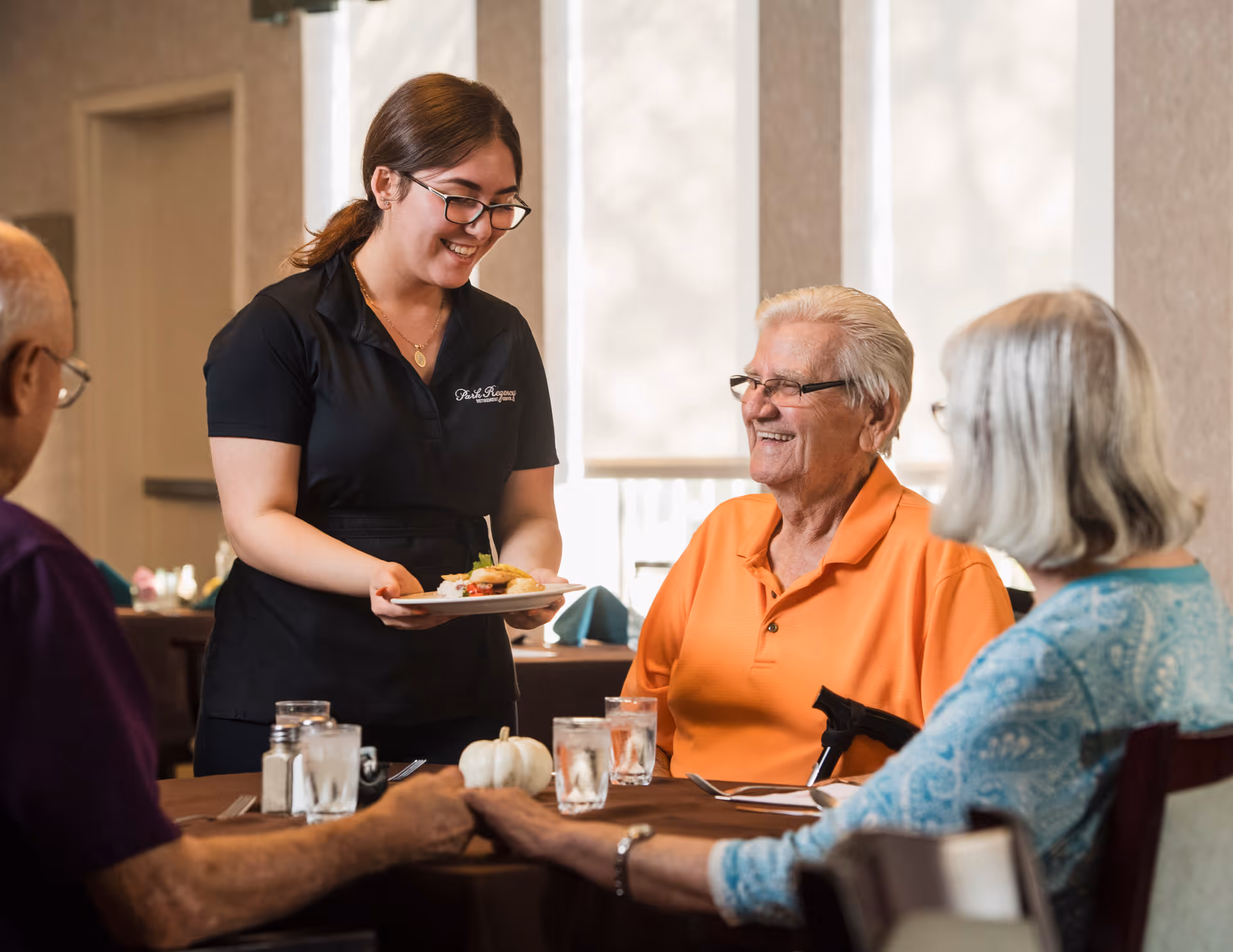 A server brings a plated meal to smiling older adults seated around a dining table in a retirement community dining room.