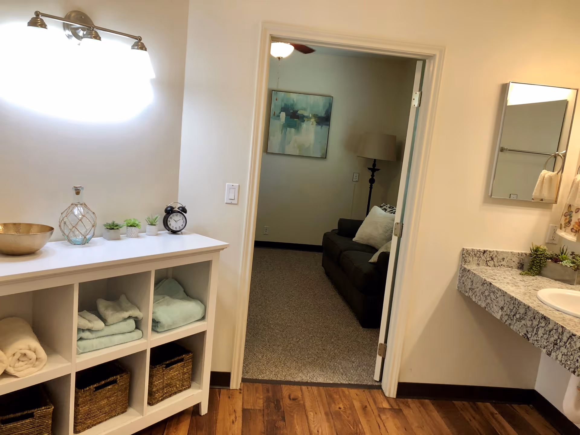 Interior view of a bathroom area with a white shelving unit holding folded towels, baskets, and decorative items. A granite countertop with a sink and a mirror is visible on the right. Through an open door, a living room area with a dark sofa, a floor lamp, and a painting on the wall can be seen.