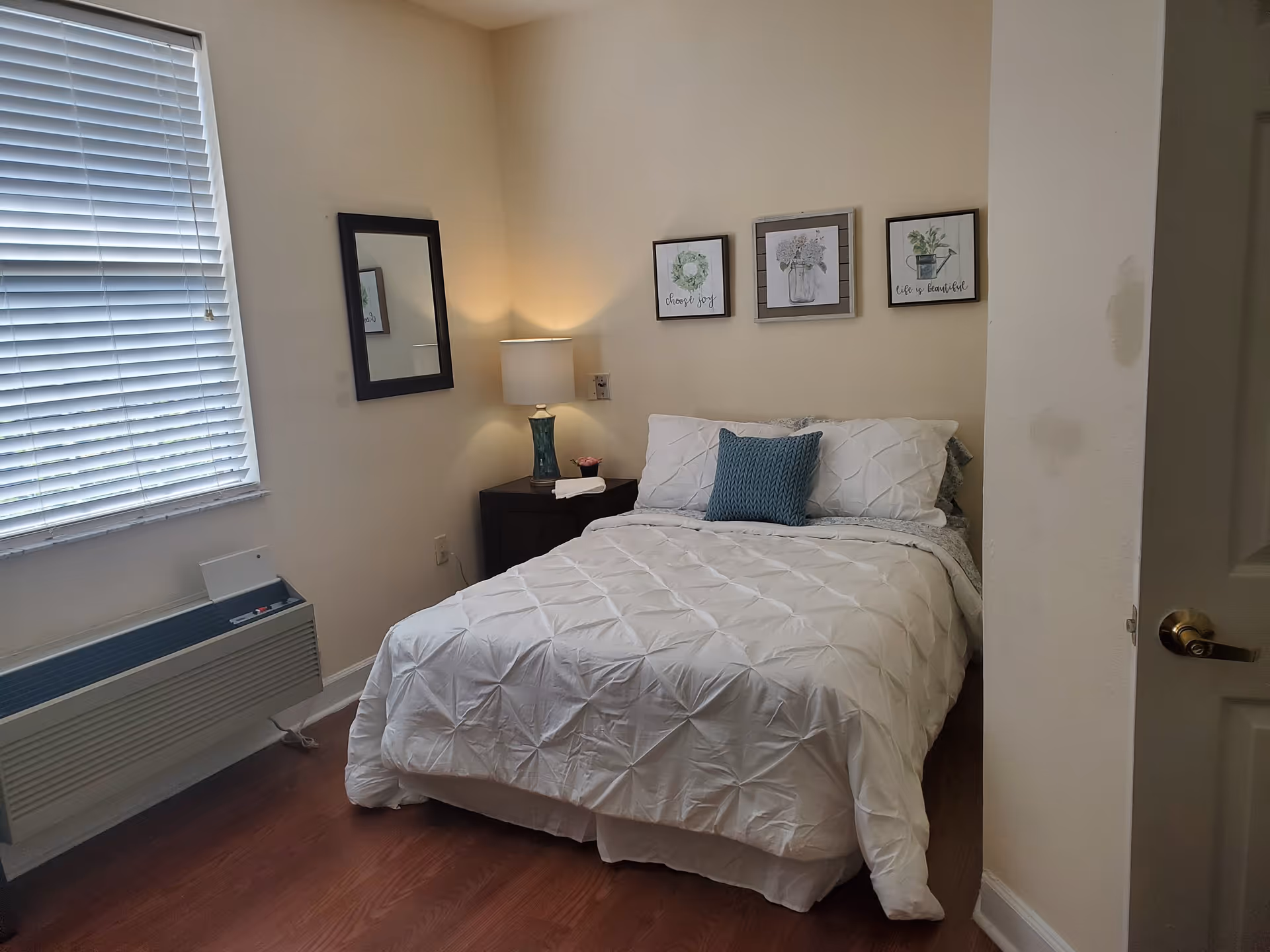 A small bedroom with a neatly made bed featuring white bedding and a blue decorative pillow. Above the bed are three framed pictures with floral and inspirational themes. To the left of the bed is a nightstand with a lamp and a small plant. A mirror hangs on the wall next to a window with closed blinds. The floor is wooden, and the walls are painted a light beige color.