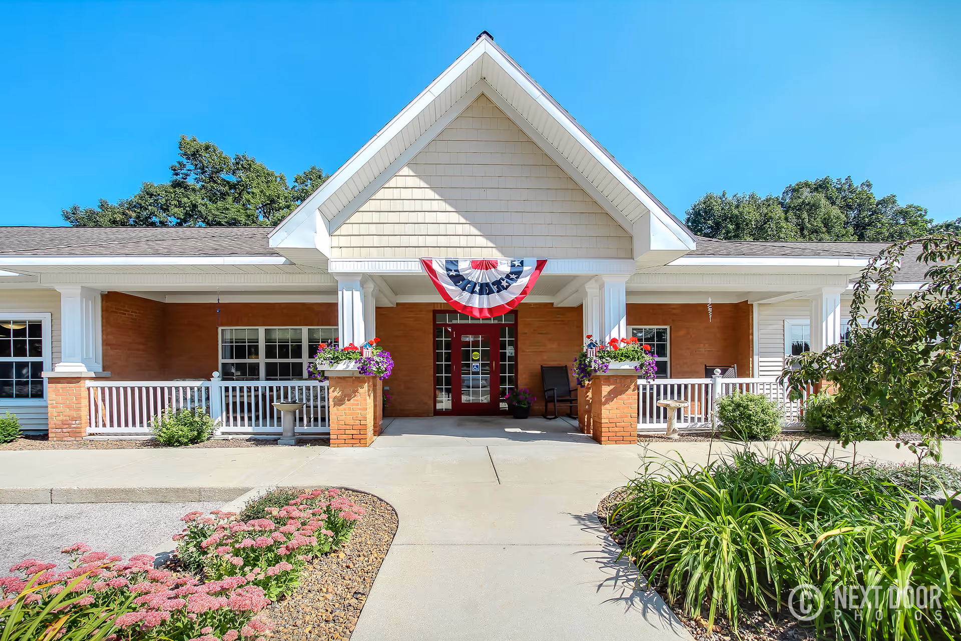 Front exterior view of Ludington Woods Assisted Living and Memory Care facility with a covered entrance, white columns, brick walls, and a patriotic red, white, and blue bunting hanging above the door. There are flower beds with pink flowers and green plants along the walkway leading to the entrance under a clear blue sky.