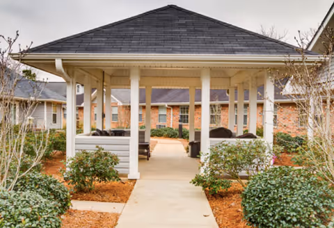 A covered outdoor pavilion with a dark shingled roof supported by white columns, surrounded by green shrubs and plants. There are benches under the pavilion and brick buildings in the background under a cloudy sky.