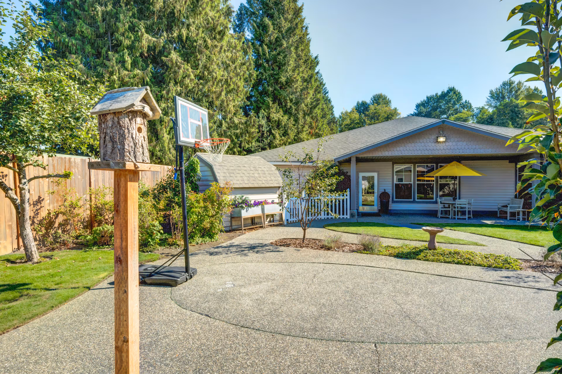 Sunny courtyard with a paved area, a birdhouse on a post, a portable basketball hoop, and a single-story building patio with a yellow umbrella.