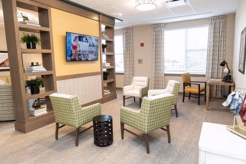 A bright and modern living room area in a senior living facility with four green plaid armchairs arranged around a small black round table. There is a wooden shelving unit with plants and books, a wall-mounted TV, large windows with patterned curtains, and a wooden desk with a chair and lamp in the corner.