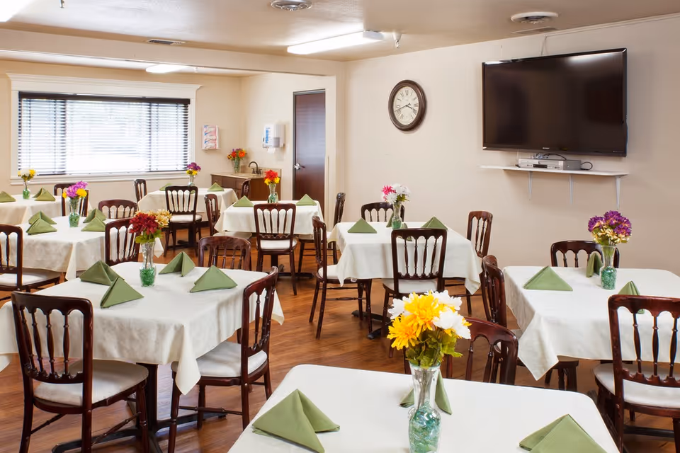 A dining room with multiple tables covered in white tablecloths, each set with green folded napkins and vases of colorful flowers. Wooden chairs surround the tables. A wall clock and a flat-screen TV mounted on the wall are visible. The room has wooden flooring and a window with blinds.