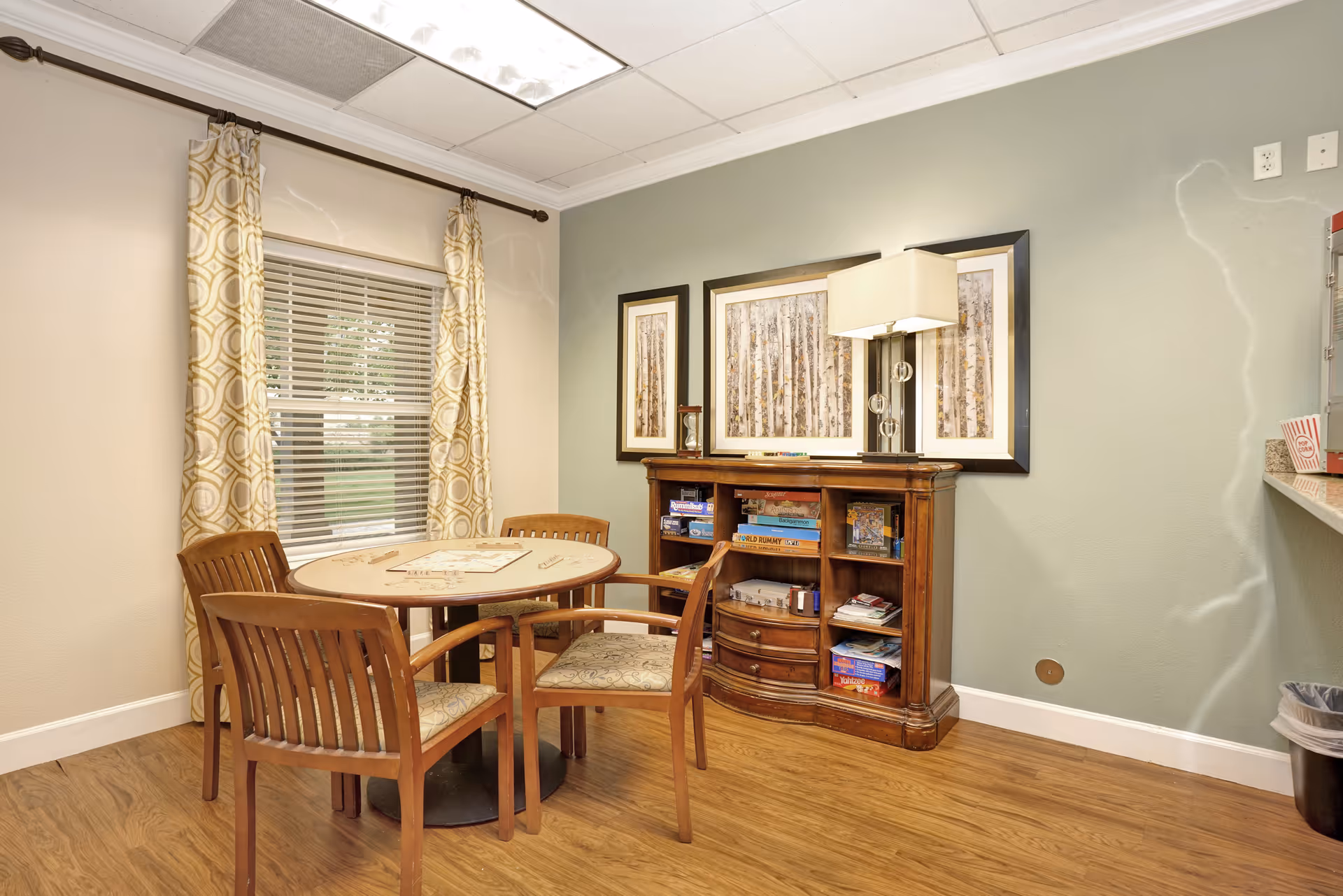 A cozy interior room with a round wooden table and four wooden chairs with patterned cushions. Behind the table is a wooden cabinet filled with board games and books. Above the cabinet are three framed pictures of birch trees and a modern table lamp. A window with patterned curtains is on the left wall, and the floor is wood.