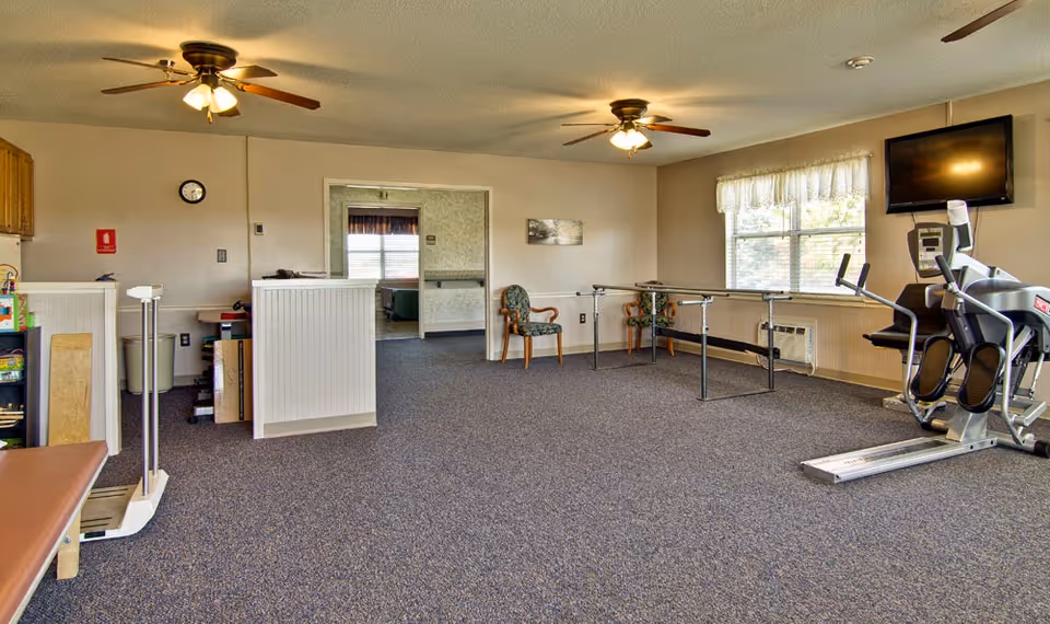 Interior room with exercise equipment including a stationary bike and parallel bars, two ceiling fans with lights, a wall-mounted TV, two chairs near a window with curtains, and a doorway leading to another room with a bed visible.