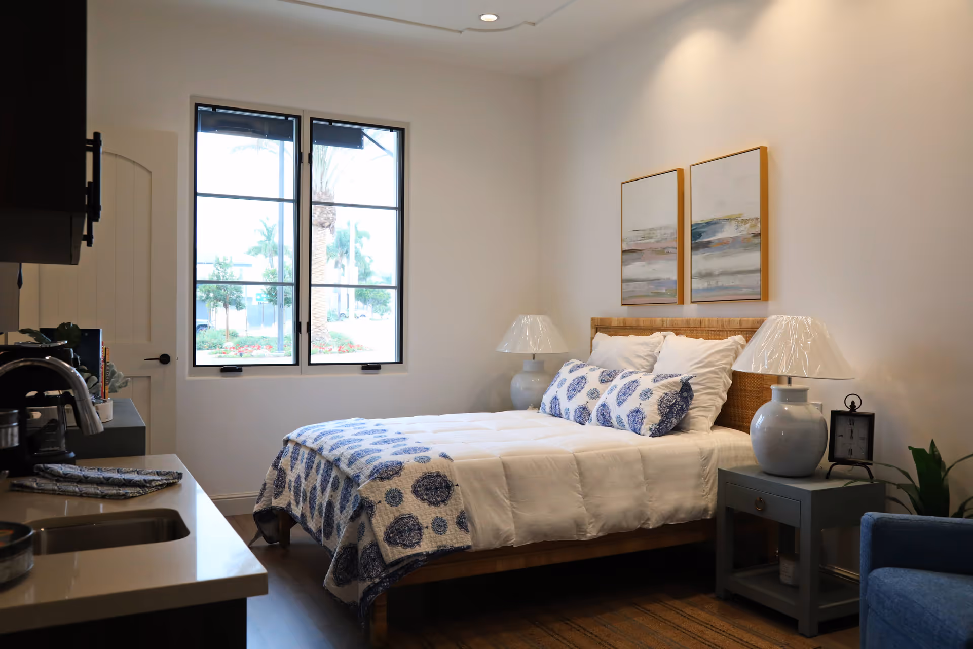 A bright bedroom with a bed dressed in white and blue patterned linens, two bedside lamps, framed artwork above the headboard, and a small kitchenette counter visible to the left.