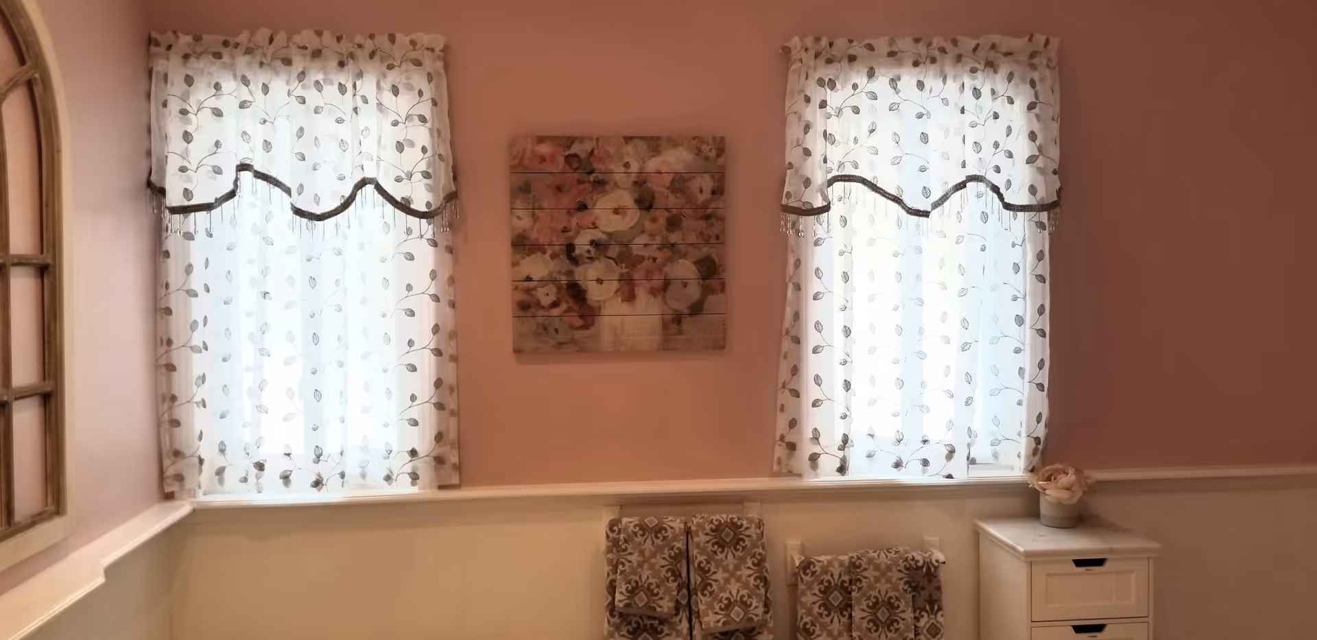 Light pink interior room with two sheer floral-curtained windows, floral wall art, patterned chairs, and a small white dresser.