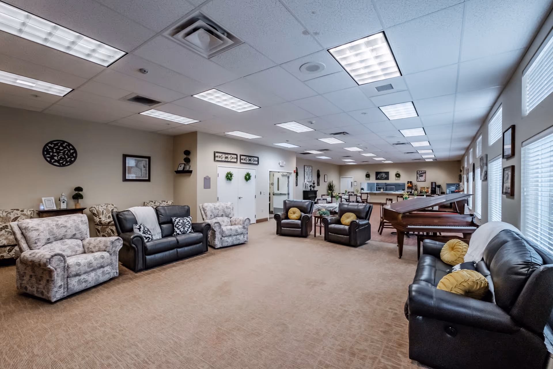A spacious, well-lit common area in a senior living facility featuring multiple armchairs and sofas arranged around the room. There is a grand piano on the right side near large windows with blinds. The walls are decorated with framed artwork and small shelves with plants. In the background, there is a dining area with tables and chairs, and a kitchen counter with appliances.